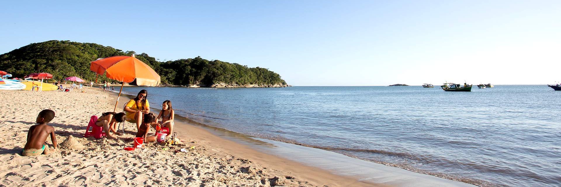 Família em um dia de sol na praia em baixo de um guarda-sol esquerda em primeiro plano, já em segundo plano outras barradas na areia, em terceiro plano pedras e vegetações. O mar a direita com algumas embarcações âncoradas