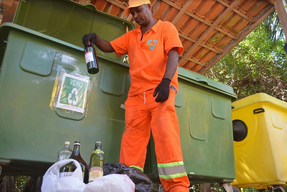 Foto de um profissional de limpeza colocando garrafas de vidro numa lixeira de coleta seletiva