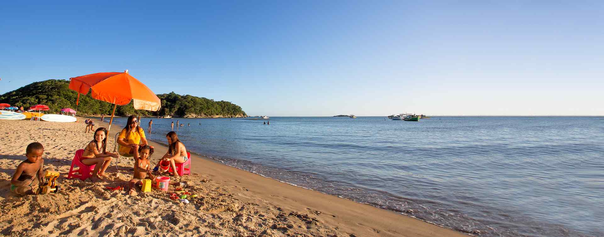 Família em um dia de sol na praia em baixo de um guarda-sol esquerda em primeiro plano, já em segundo plano outras barradas na areia, em terceiro plano pedras e vegetações. O mar a direita com algumas embarcações âncoradas