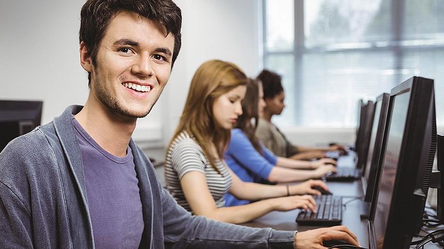 Foto no primeiro plano de um jovem sorrindo e no segundo plano três jovens usando computadores durante um curso de informática