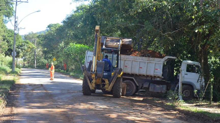 Foto de retroescavadeira recolhendo entulhos e ao fundo operários realizando capina