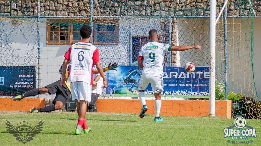 Foto de um lance da partida onde o jogador está chutando a bola em direção ao gol, e o goleiro pulando para tentar defender