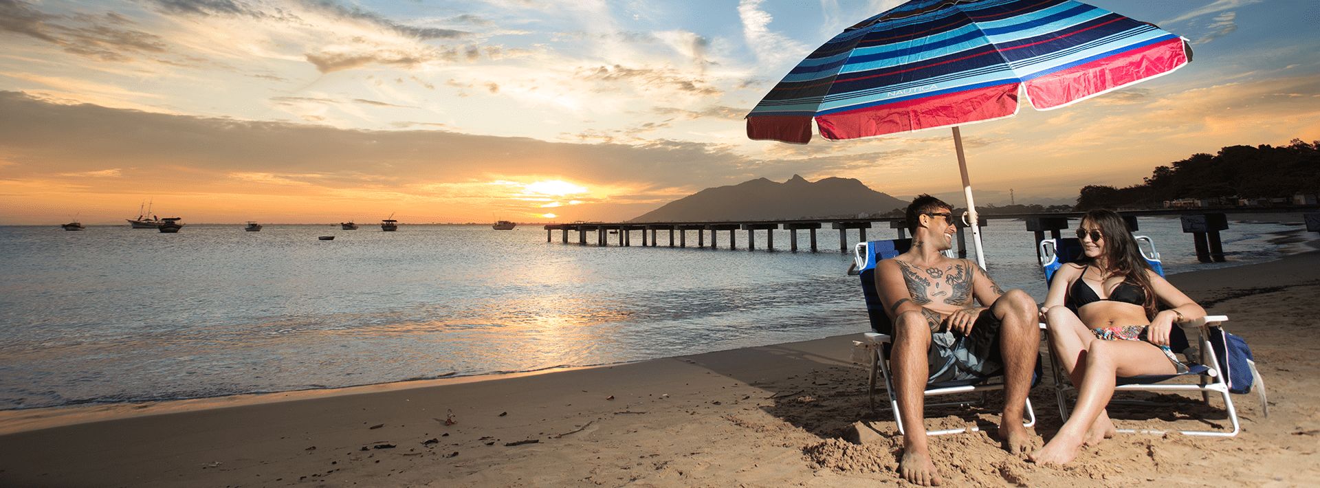 Família em um dia de sol na praia em baixo de um guarda-sol esquerda em primeiro plano, já em segundo plano outras barradas na areia, em terceiro plano pedras e vegetações. O mar a direita com algumas embarcações âncoradas