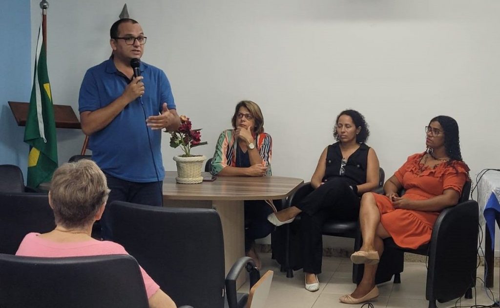 Secretário de Educação com microfone na mão ao lado de três conselheiros sentados