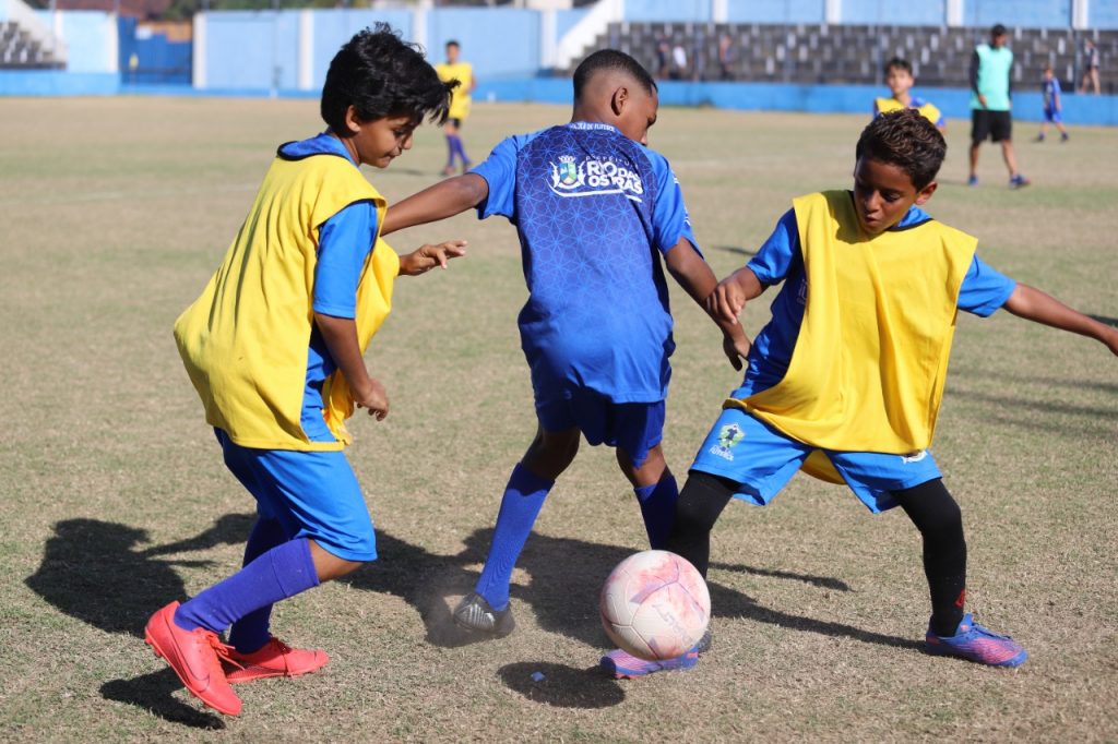 Três jogadores da categoria infantil disputam a bola no gramado do campo