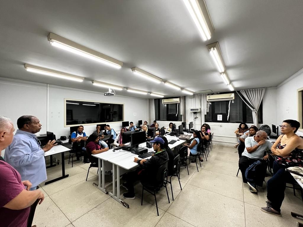 Alunos sentados em frente aos computadores recebem informações do professor que está em pé