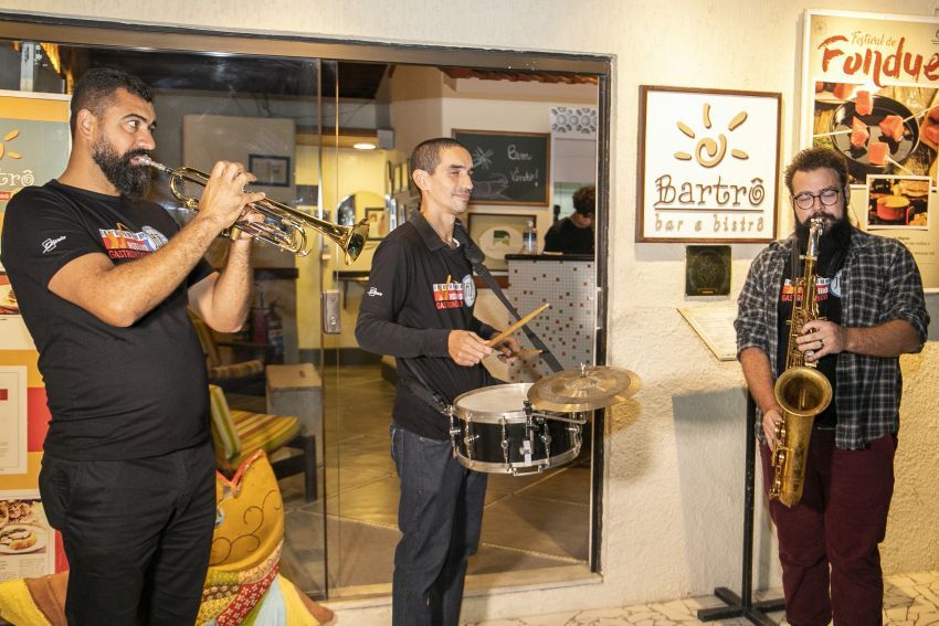 Trio de músicos tocando. à esquerda, músico tocando trompete. Ao centro tocando caixa. à direita, tocando sxofone.