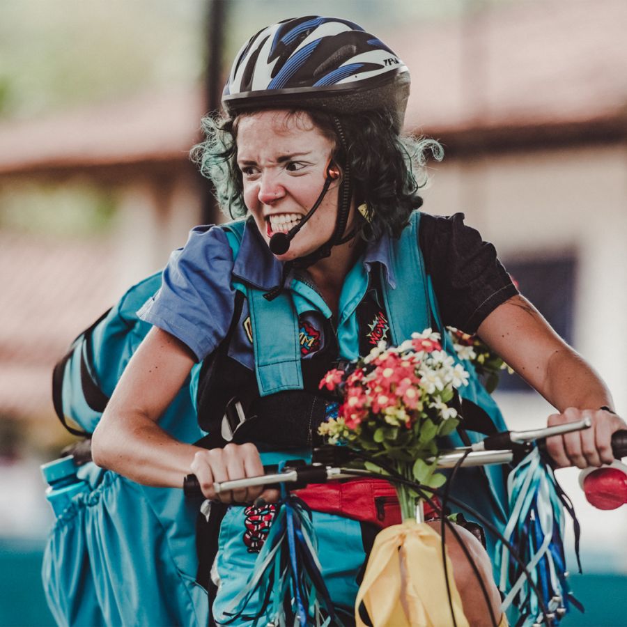 Caracterizada como palhaça, com maquiagem e capacete, atriz anda em uma bicicleta