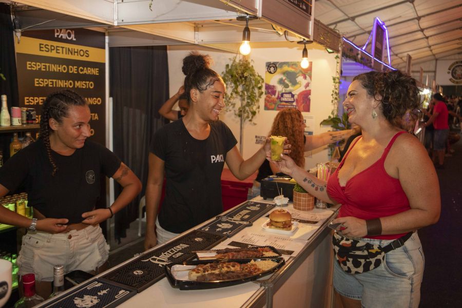 Duas mulheres jovens sorridentes, atrás do balcão, servem drinque para uma mulher também jovem e sorridente