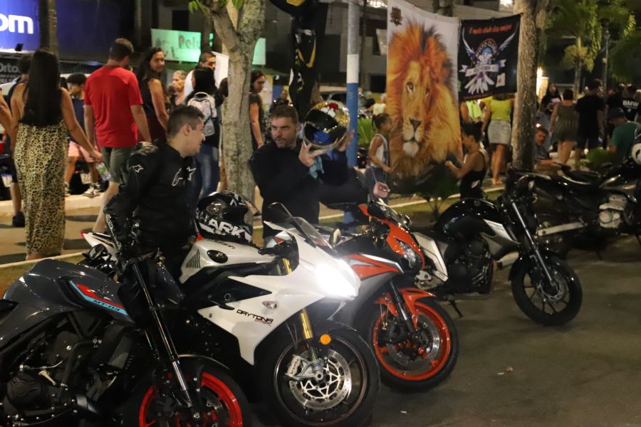 Na frente da foto, dois homens ao lado de motos conversam; um deles está com o capacete nas mãos. Aos fundos, bandeiras de motoclubes e pessoas que caminham no evento