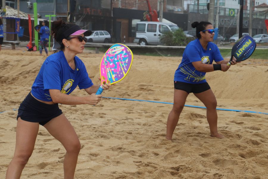 Na foto, duas atletas estão na areia da praia, cada uma segura uma raquete de tenis de praia. Ambas usam camisa azul e estão de oculos escuros.