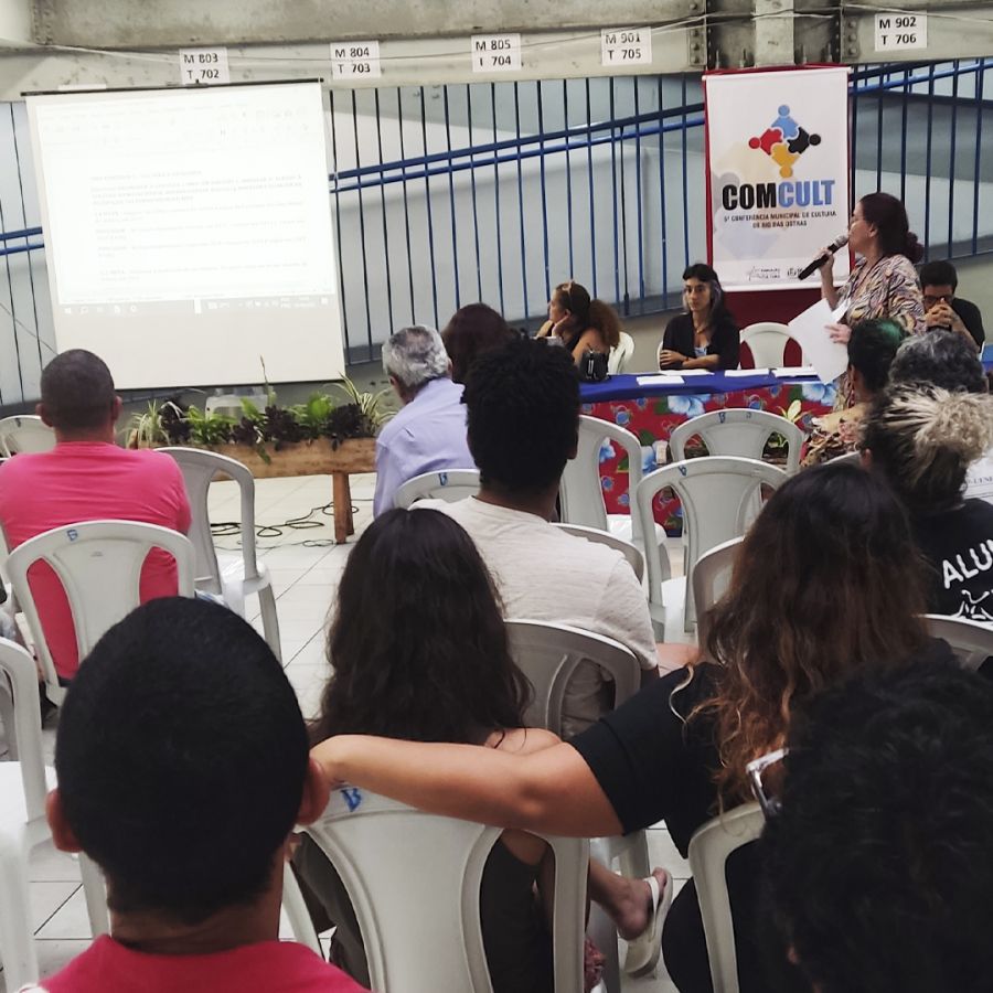 Mulher de pé, ao microfone, com banner da Conferência de Cultura aos fundos. Pessoas acompanham a explicação dela, sentadas em cadeiras