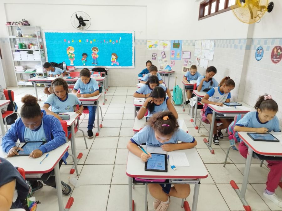 Sala de aula com alunos sentados um atrás do outro, em fileiras, e usando tablets