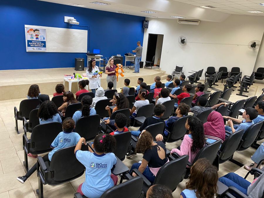Auditório de uma escola com dois profissionais de saúde bucal em pé palestrando para crianças sobre cuidados em saúde bucal