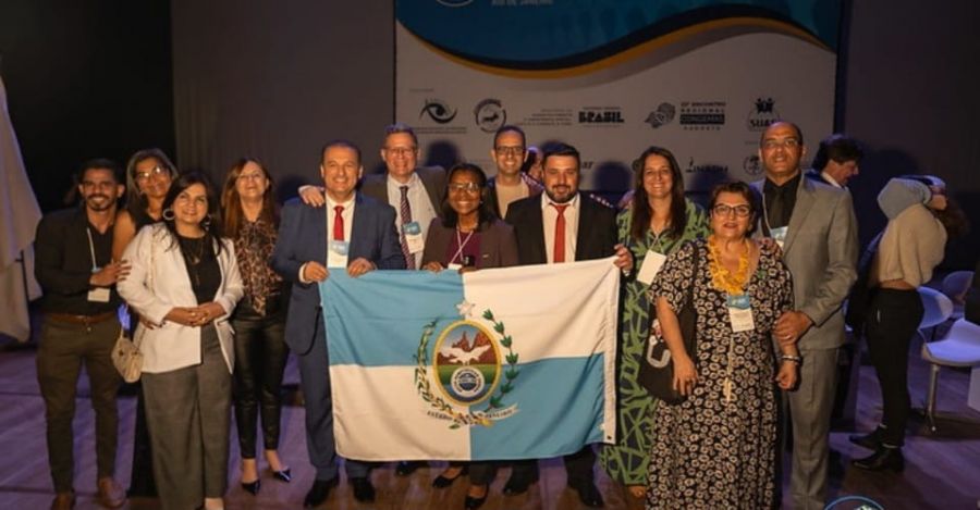Representantes de assistências sociais municipais em pé segurando a bandeira do Estado do Rio de Janeiro.