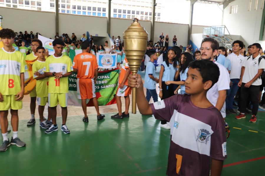 Aluno adolescente de pele morena usando uniforme esportivo segurado uma taça. Outros estudantes observado.
