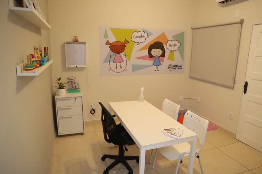 Sala com mesa branca e cadeiras, quadro colorido na parede, brinquedos nas prateleiras
