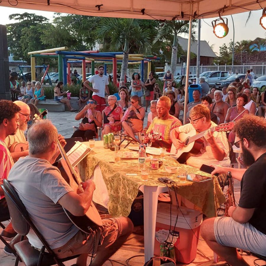 Músicos reunidos ao redor da mesa, sentados, tocando instrumentos. Público, também sentado, observa a apresentação