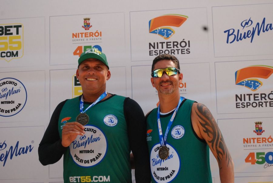 Na foto, uma dupla de homens posa segurando a medalha de 3º lugar do campeonato de volei de praia.