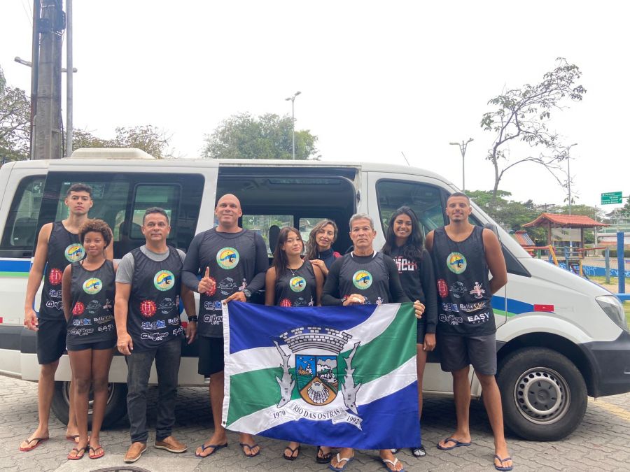 Na foto os atletas de volei de praia posam segurando a bandeira de Rio das Ostras em frente a van do Esporte.