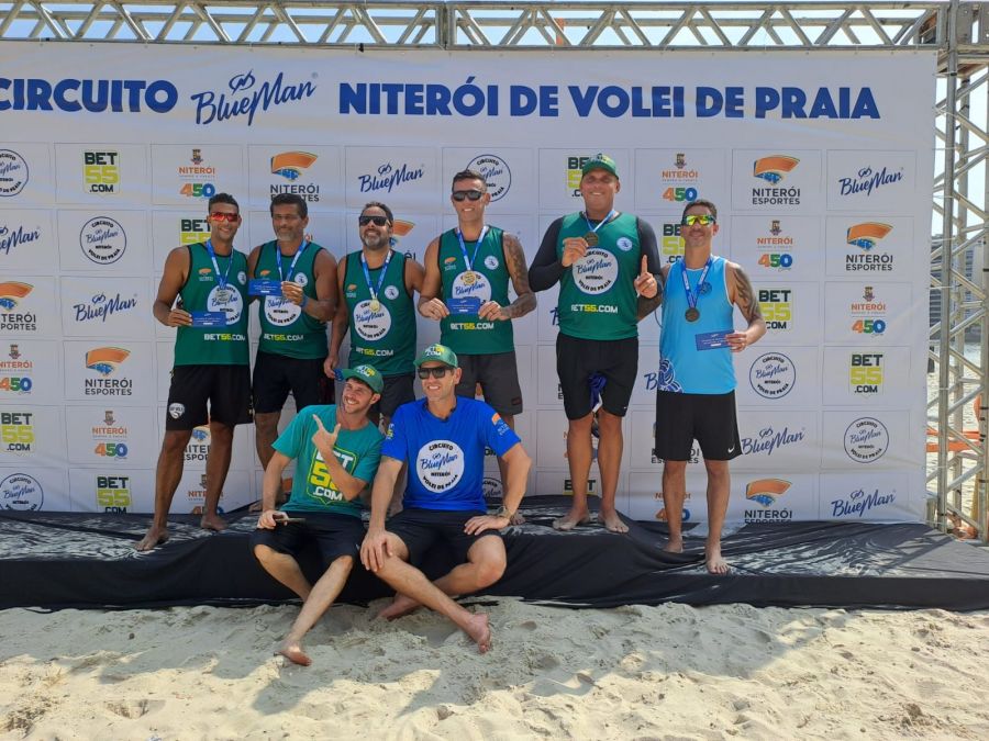 Na foto, 08 atletas posam segurando a bandeira de Rio das Ostras em frente a um banner do campeonato de volei de praia de Niterói.