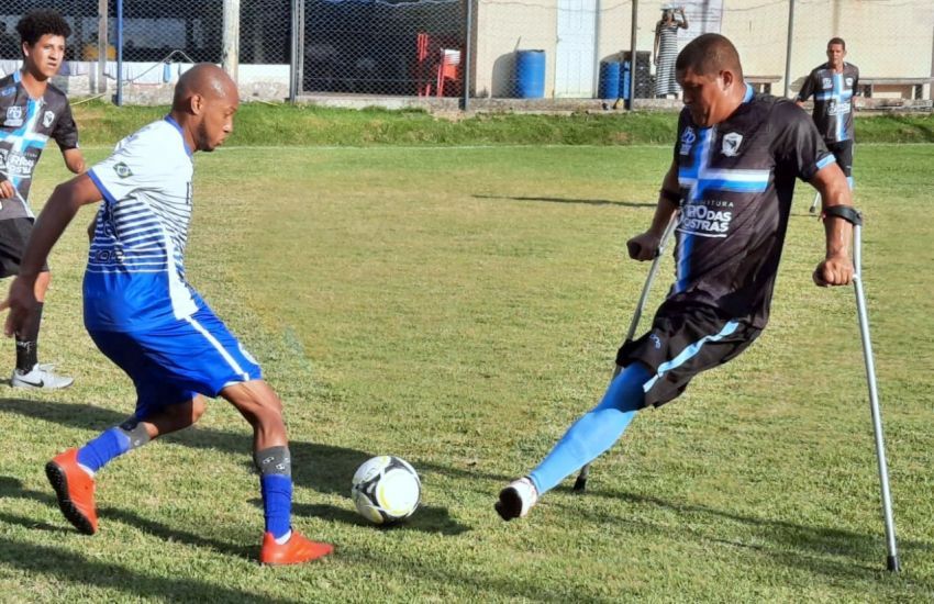 Dois jodores de futebol. O atleta da direita é deficiente e está de muleta. Estão em um campo de futebol.