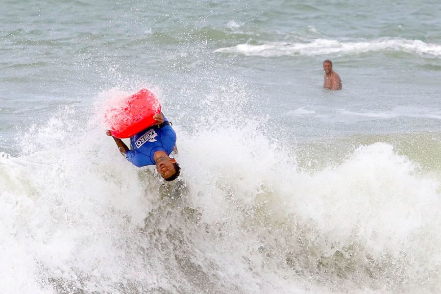 Na foto, um atleta de bodyboard faz uma manobra na água e fica de cabeça pra baixo em uma onda