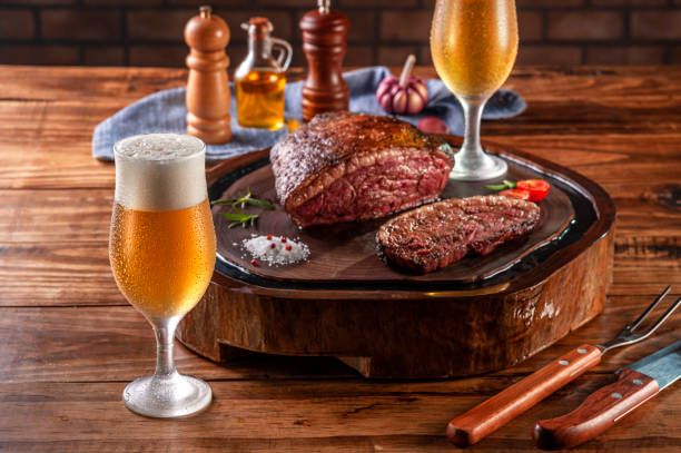 Na foto, dois copos de cerveja e uma tabua com churrasco aparecem em primeiro plano. No fundo, uma bandeija com temperos compõe a imagem.