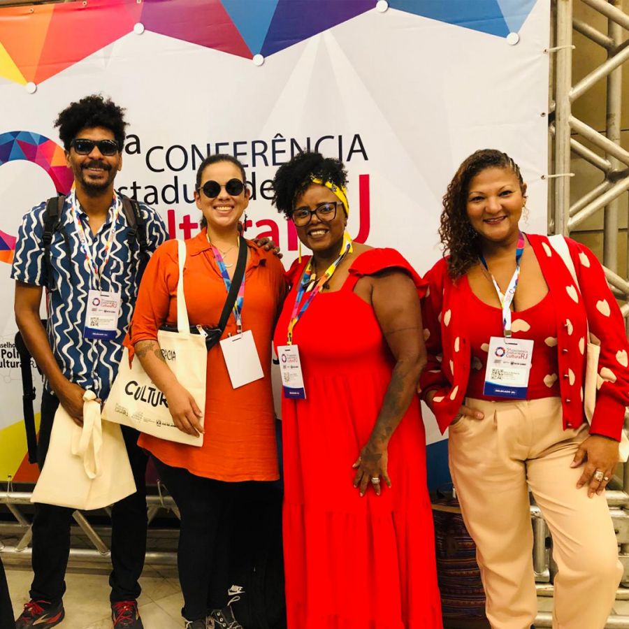 Quatro pessoas, um homem negro, uma mulher branca, uma mulher negra e uma mulher parda pousam em frente ao banner da Conferência Estadual de Cultura