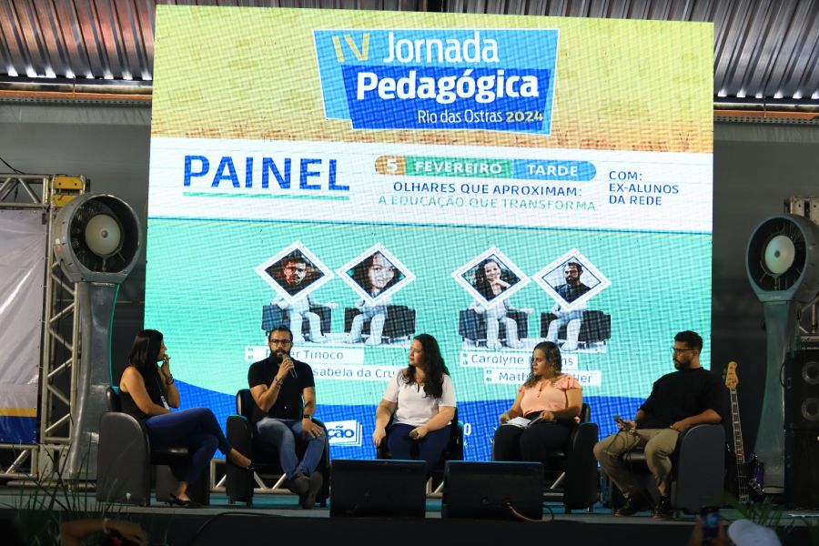 Cinco jovens, dois homens e três mulheres, sentados em cadeiras no palco, tendo aos fundos painel de LED da IV Jornada Pedagógica anunciando o tema do painel 