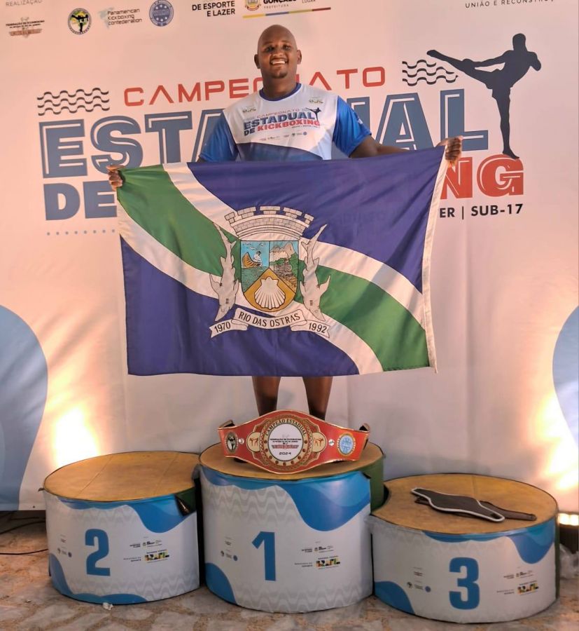 Na foto, o atleta Alan aparece no local mais alto do pódio com o cinturão de campeão e segurando a bandeira de Rio das Ostras